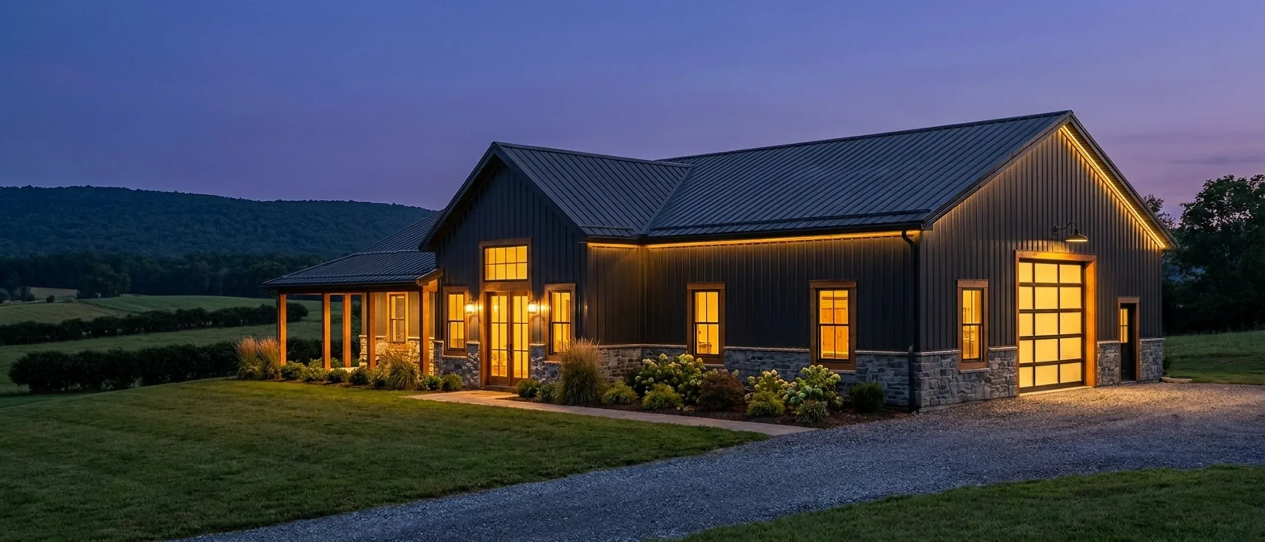 A barndominium at dusk with warm light glowing from every window and exterior lighting along the porch and roofline, a long forested ridge dark on the horizon.